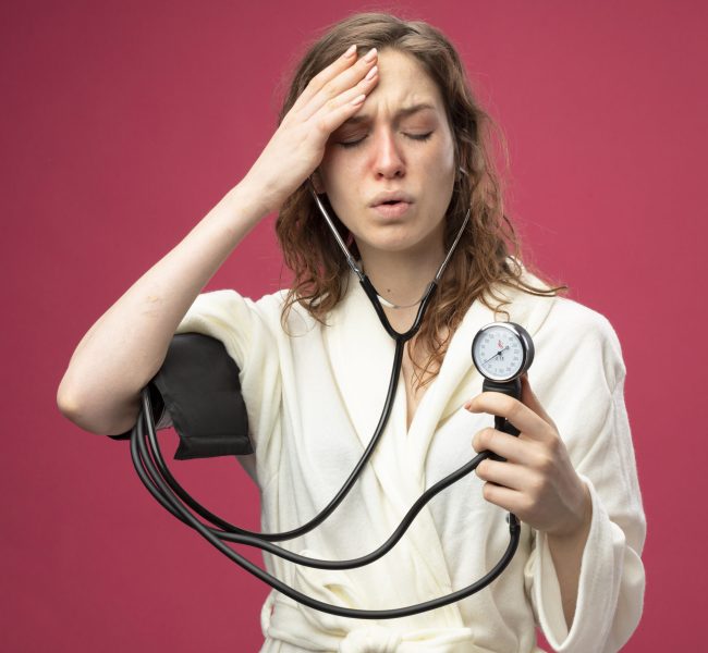 weak young ill girl with closed eyes wearing white robe measuring her own pressure with sphygmomanometer putting hand on forehead isolated on pink background